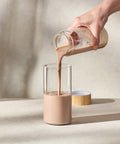 Person pouring a creamy liquid from a glass jar into another glass on a neutral background