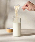 Person adding Juice Plus+ Complete powder to a glass bottle of milk on a neutral background
