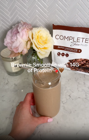 Hand holding a brownie smoothie with a package of Complete Protein in the background.