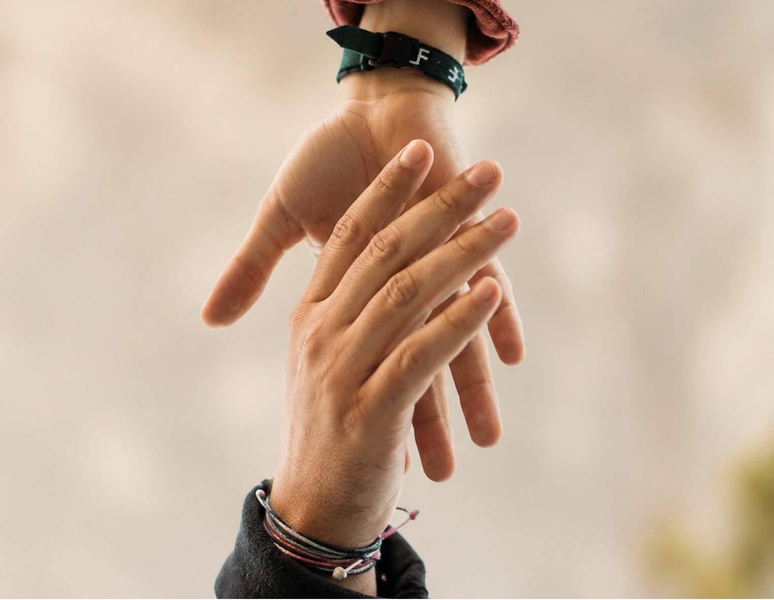 Two hands gently touching, each wearing a thin black bracelet. The background is a soft, out-of-focus beige tone.
